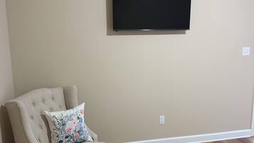 A living room with a beige wall, a flat-screen TV mounted above a beige armchair with a floral pillow, and a wooden floor.