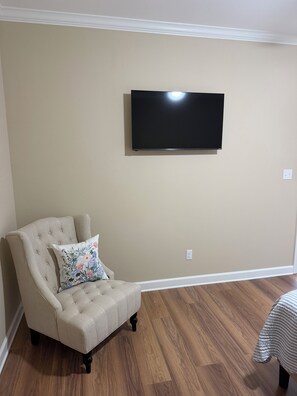 Una sala de estar con pared beige, un televisor de pantalla plana montado sobre un sillón beige con un cojín floral, y suelo de madera.