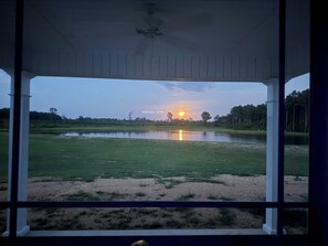 A view from a covered porch overlooking a grassy field with a body of water and trees in the distance.