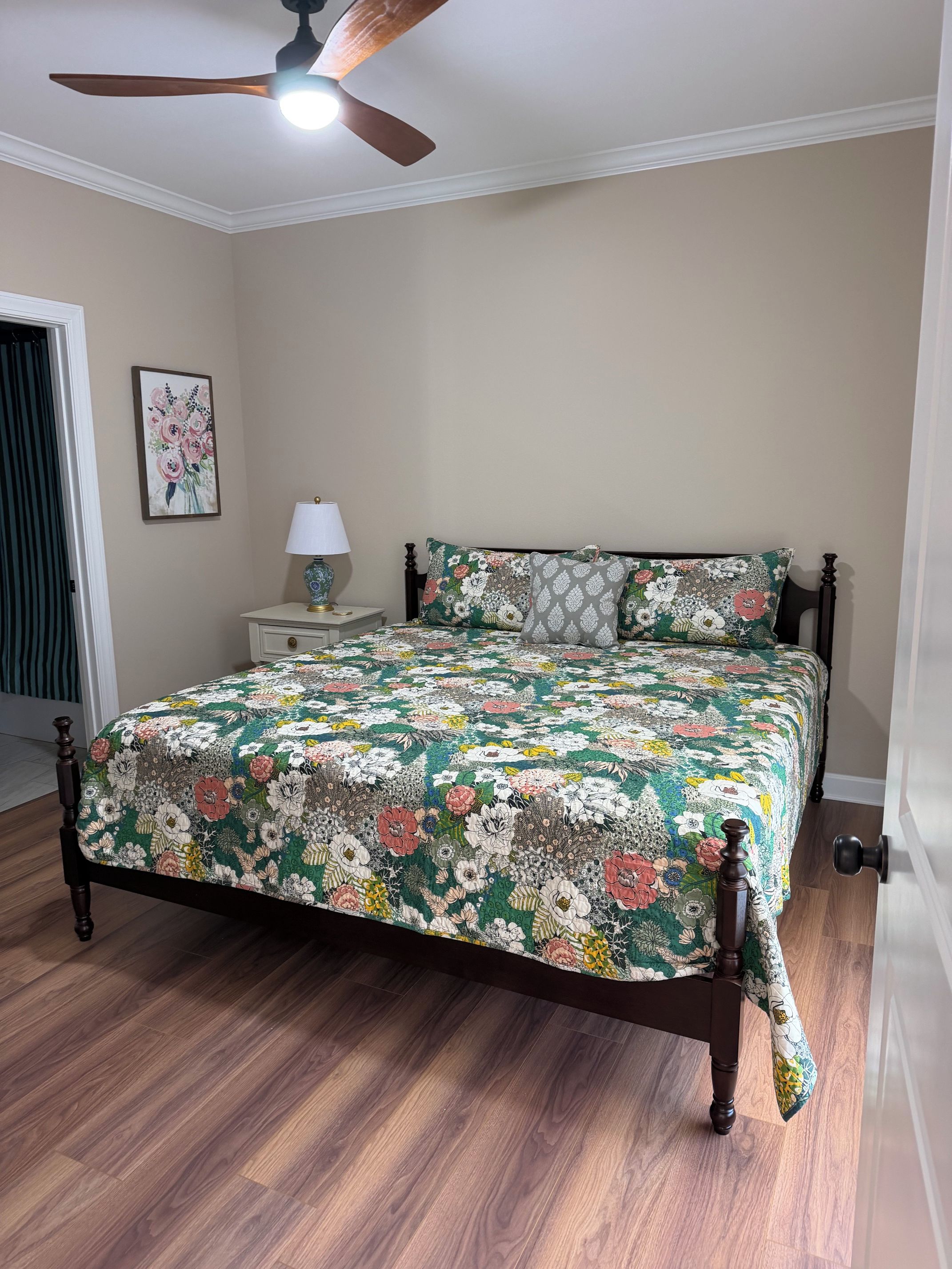 A bedroom with a wooden bed, a ceiling fan, a nightstand with a lamp, and a floral wall art piece.