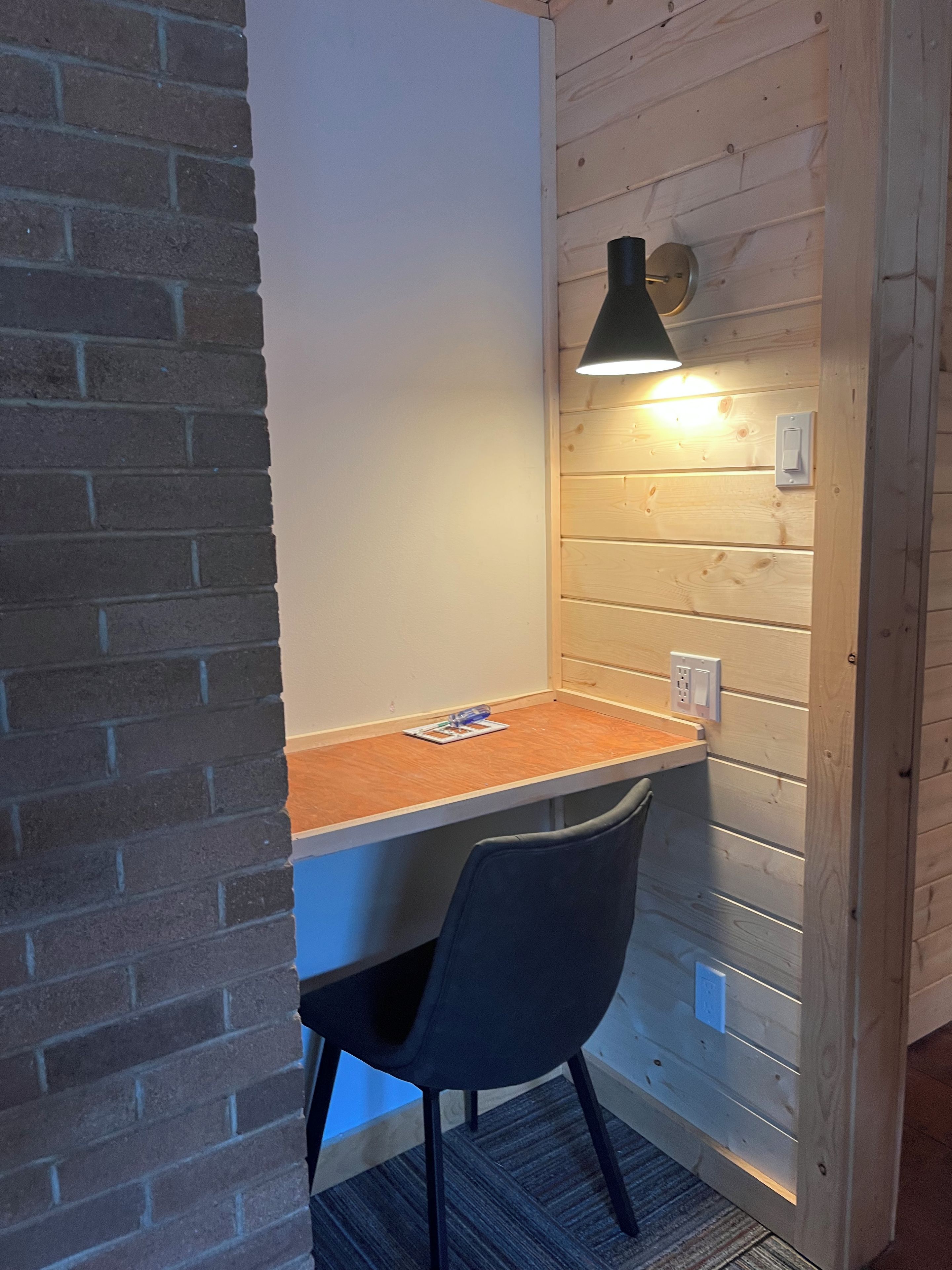 A corner room with a brick wall, a wooden desk, a chair, and a wall-mounted lamp.
