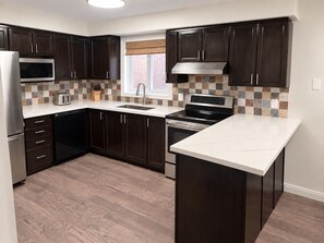 Una cucina moderna con mobili in legno scuro, un forno a microonde in acciaio inossidabile, un forno integrato e una grande isola con piano di lavoro bianco.