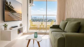 A modern living room with a green sofa, a flat-screen TV, a small table with a vase, and a balcony with yellow chairs.