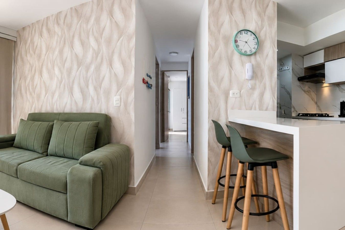 Un espace de vie moderne avec un canapé vert, un coin cuisine doté d’un pare-feu en marbre et un couloir menant à une autre pièce.