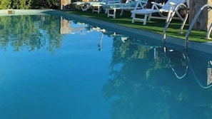 Una piscina con agua azul cristalina, rodeada de sillas reclinables blancas y una zona verde.