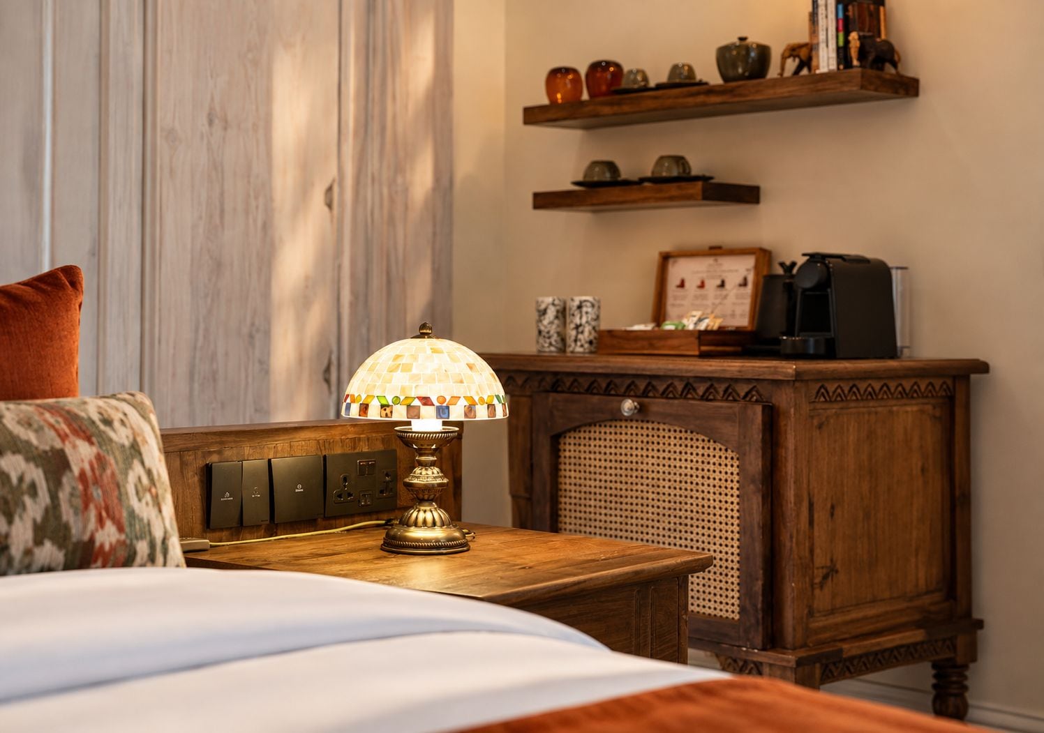 A wooden bedside table with a lamp, a bed with a patterned pillow, and a wooden cabinet with shelves above.