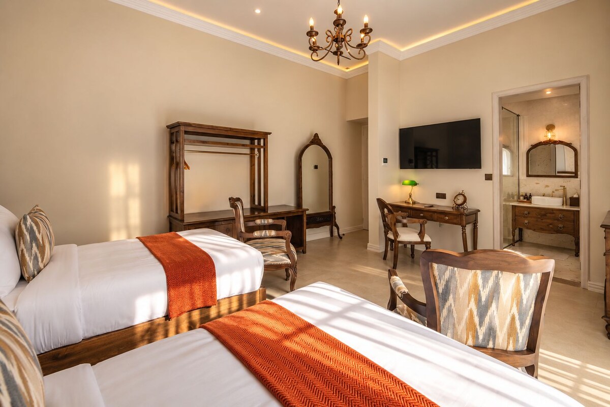 A hotel room with two beds, a desk, a chair, a television, a mirror, and a bathroom with a sink and mirror.