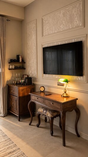 A room with a desk, a chair, a shelf with books and decorative items, a clock, and a wall-mounted television.