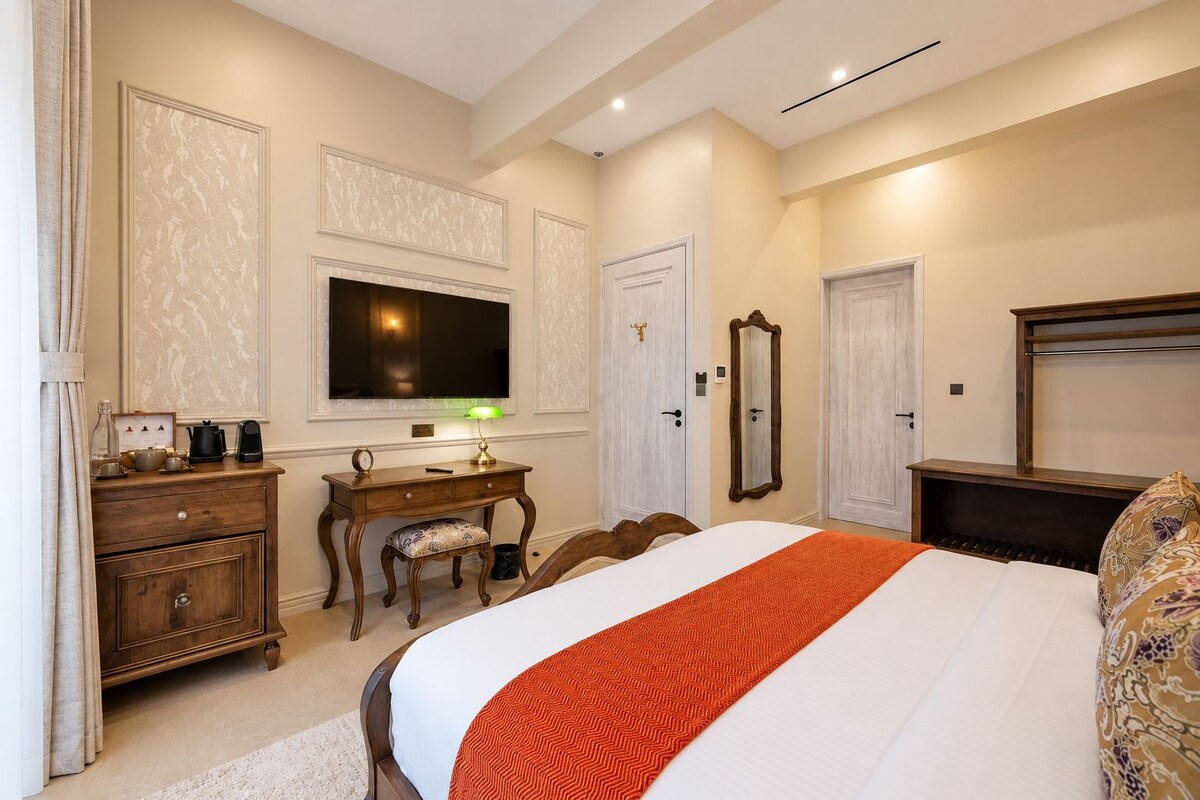 A hotel room with a bed, a desk with a lamp, a television, a mirror, and a piano.