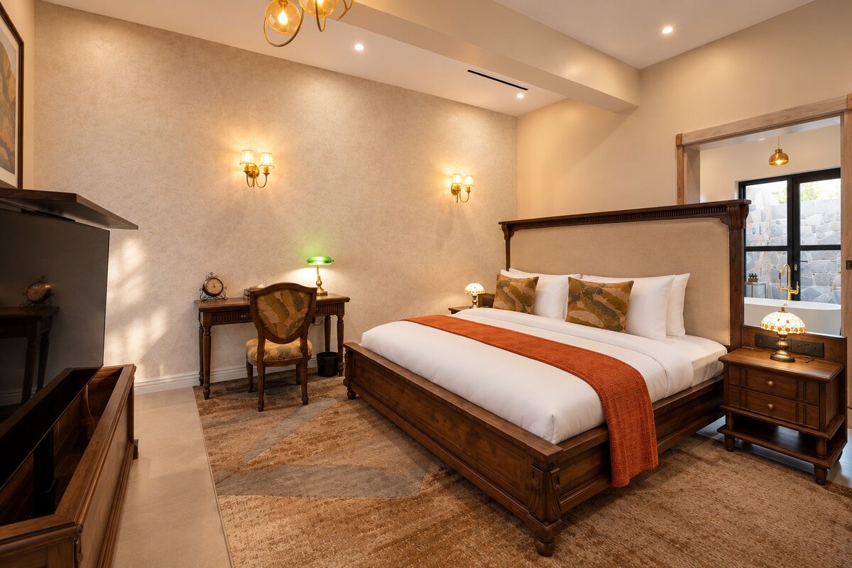 A hotel room with a large bed, a desk, a chair, a small table with a lamp, and a window with a view.