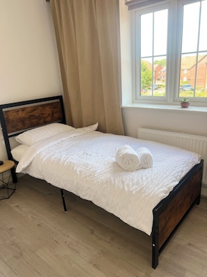 Un lit bien fait, avec du linge de lit blanc et une serviette enroulée, une tête de lit en bois, une petite table de chevet et une fenêtre donnant sur un quartier résidentiel.