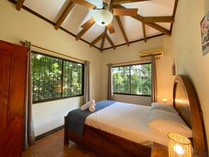 Quarto com teto de madeira, duas janelas com grades de metal, cama com roupa de cama branca e uma luminária de cabeceira.