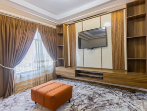 Ein Zimmer mit einem wandmontierten Flachbildfernseher an einer Holzwand, einem gepolsterten Hocker in der Mitte und einem großen Fenster mit Vorhängen.
