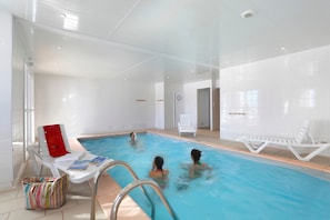 Ein überdachter Swimmingpool mit klarem Wasser, umgeben von weißen Wänden und einem gefliesten Boden. Liegestühle und ein Tisch mit einer gestreiften Tasche darauf sind vorhanden.