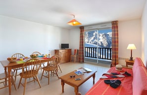 Ein Zimmer mit Esstisch, Stühlen, Fernseher, Sofa, einem Fenster mit Bergblick und einem Balkongeländer.