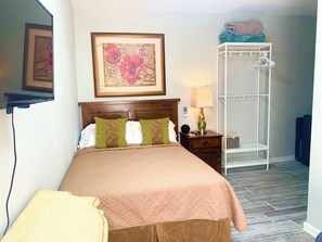 Un lit bien fait, recouvert d’une couvre-lit à motifs, une lampe de chevet, un tableau encadré accroché au mur et un meuble de rangement blanc avec des cintres.