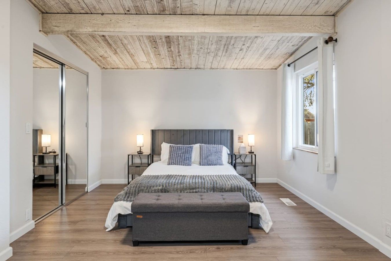 A bedroom with a wooden ceiling, a large bed with a grey upholstered bench, two bedside tables with lamps, and a large window on the right.