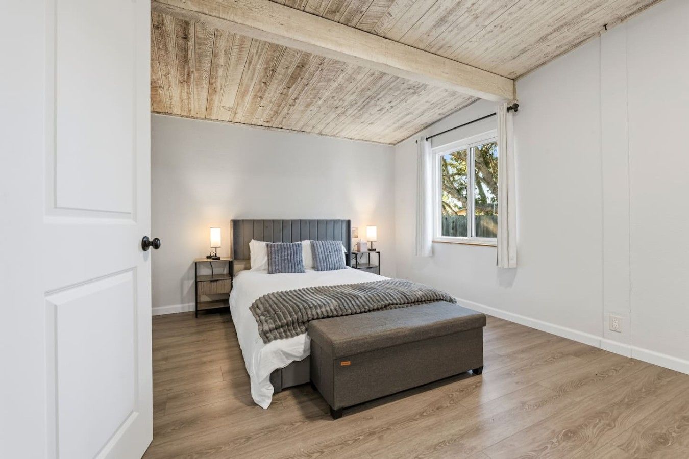 A bedroom with a wooden ceiling, a large bed, bedside tables, and a window with a view.