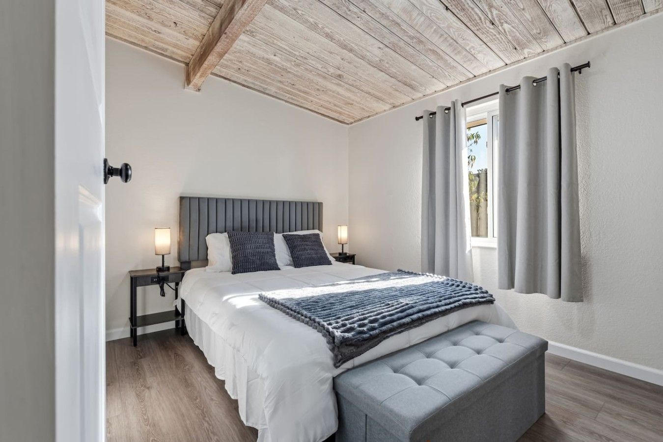 Un dormitorio con cama, lámparas de noche, un banco y una ventana con cortinas.