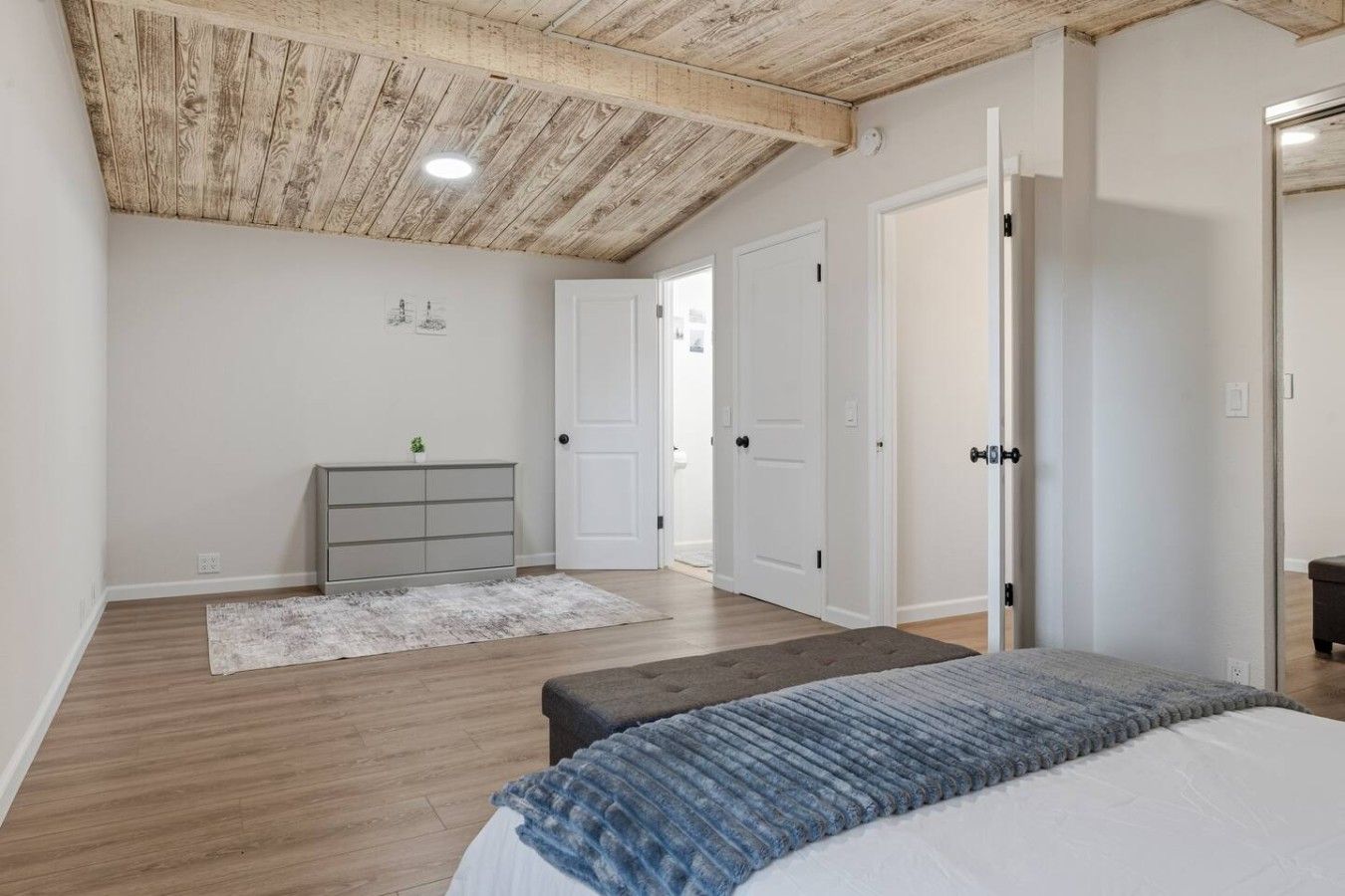 A bedroom with a wooden ceiling, a bed with a blue blanket, a grey dresser, and white doors.