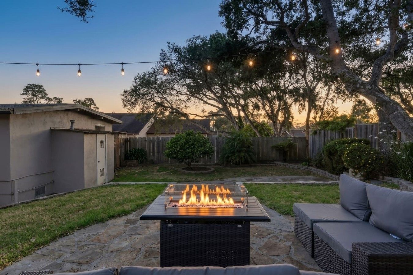 A backyard with a fire pit, outdoor furniture, and string lights.