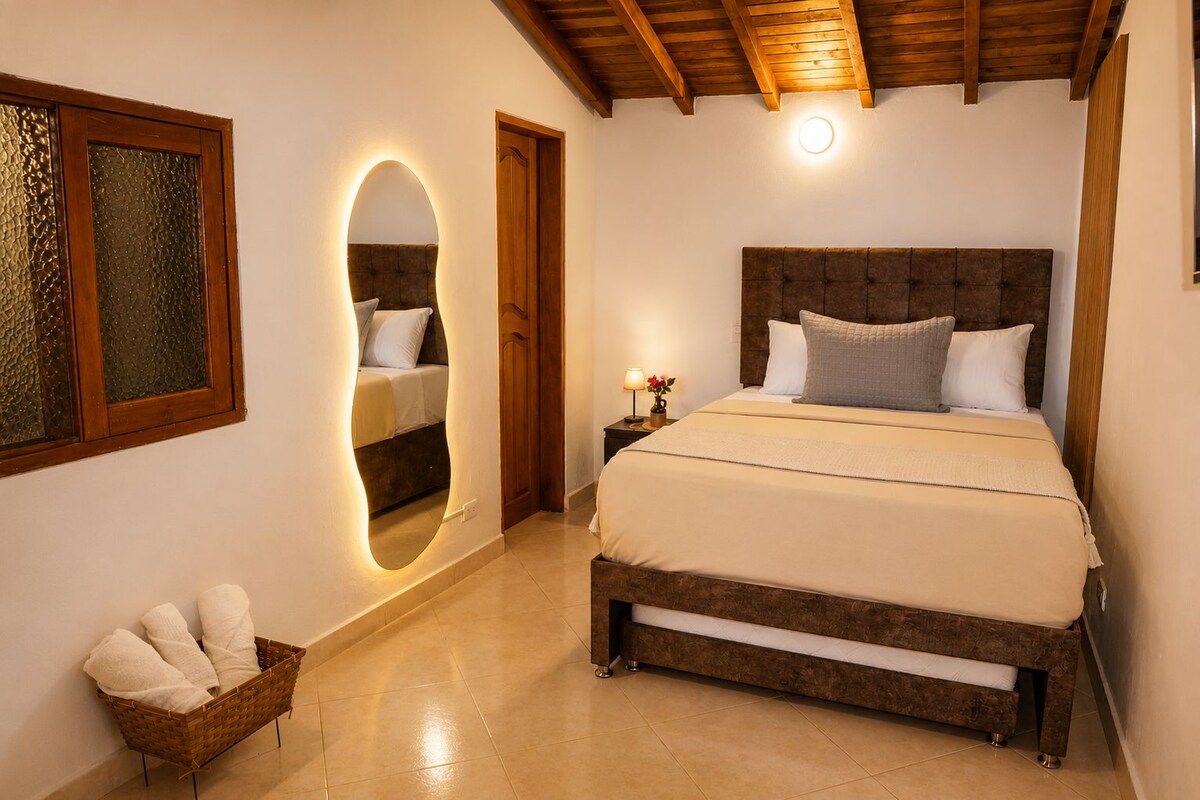 A bedroom with a large bed, a wooden headboard, a nightstand with a lamp, a mirror, and a wicker basket with towels.