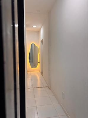 A hallway with white walls and a tiled floor leading to a room with a curved mirror. - Primos 2 Suites & Hostel (Búzios)