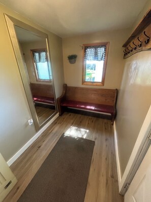 A narrow hallway with a wooden bench, a small window with checkered curtains, and a mirror on the left wall.