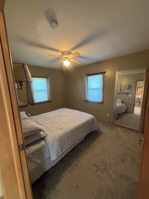 A bedroom with a bed, a ceiling fan, two windows, and a door leading to another room.
