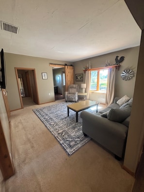 Ein Wohnzimmer mit Sofa, Sessel, Couchtisch und einem gemusterten Teppich. Eine Tür führt in einen anderen Raum, ein Fenster mit Vorhängen und eine an der Wand befestigte Uhr sind ebenfalls zu sehen.