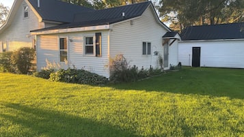 Une maison blanche d’un seul étage, avec un toit foncé, un porche à l’avant et un petit jardin devant, avec de la pelouse et des arbustes.