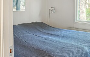 Un lit simple recouvert d’une couverture bleue à motifs, dans une pièce dotée d’une fenêtre et d’une lampe fixée au mur.