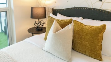Un lit avec une literie blanche et deux oreillers, un blanc et un doré, dans une pièce dont un mur est orné d’un motif géométrique et où se trouve une lampe sur une petite table placée à côté du lit.