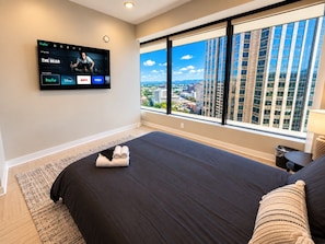 A hotel room with a large bed, a TV mounted on the wall, and a view of the city through large windows. - Birmingham Hideaway | 2BR + King (Birmingham)