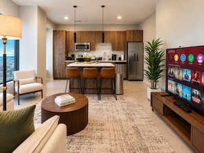 A modern living room with a flat-screen TV, a coffee table, and a sofa. In the background, there is a kitchen area with wooden cabinets, a microwave, and a refrigerator. - Birmingham Hideaway | 2BR + King (Birmingham)