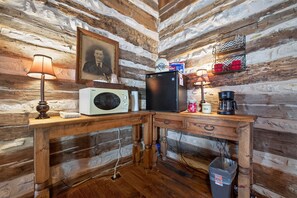 A rustic cabin interior with a wooden table, a microwave, a refrigerator, a coffee maker, and a shelf with red mugs. - Cabin w/King bed on Wine Trail, Walk to Wineries! (Fredericksburg)