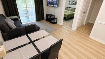 Una sala de estar moderna con un sofá, una mesa de comedor con sillas, un televisor de pantalla plana montado en la pared y vistas al exterior a través de un ventanal grande.
