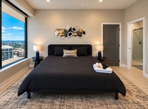 Quarto moderno com uma cama grande, duas mesas de cabeceira, uma obra de arte na parede e vista da cidade por uma ampla janela.
