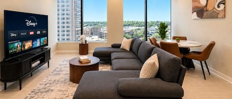 A modern living room with a sectional sofa, a flat-screen TV, a dining area, and large windows offering a city view.