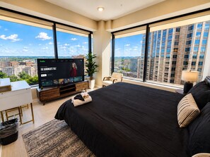 A modern hotel room with a large bed, a television, a desk, and a view of the city. - The Birmingham View | Spacious 2BR (Birmingham)