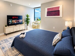 Une chambre moderne avec un grand lit, un téléviseur diffusant Netflix, une vue sur la ville et un décor minimaliste.