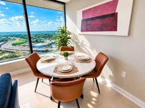 A dining area with a round table, brown chairs, and a view of a cityscape through large windows. - The City Glow | Spacious 2BR | 2 Queens (Birmingham)
