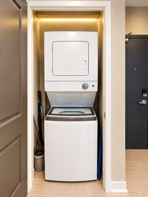A compact, modern laundry room with a built-in washer and dryer, a small trash bin, and a door leading to another room. - Downtown View Retreat | 3BR | 2 Kings (Birmingham)