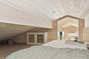 A bedroom with a sloped ceiling, wooden walls, a bed with a white duvet, a small table with a vase and mugs, and a window with a view of greenery outside. - Cosy, modern holiday home in a lovely area (Juelsminde)