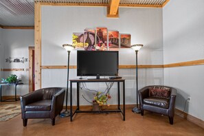 Ein Wohnzimmer mit einem Flachbildfernseher auf einem Holzbodenständer, zwei Ledersesseln, einem kleinen Tisch mit Blumenvase und einem an der Wand hängenden Bild über dem Fernseher.