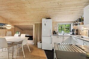 A modern kitchen with a built-in refrigerator, a dining area with a table and chairs, and a window with a view of greenery. - Lovely house on Helgenæs (Knebel)