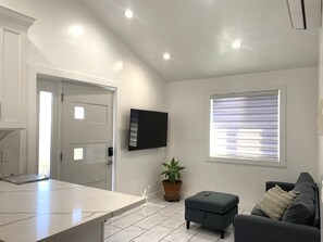 A modern living room with a flat-screen TV mounted on the wall, a grey sofa, a small ottoman, and a potted plant. - Modern Comfort Meets Luxury — Your Stylish North Hollywood Getaway (NORTH HOLLYWOOD)