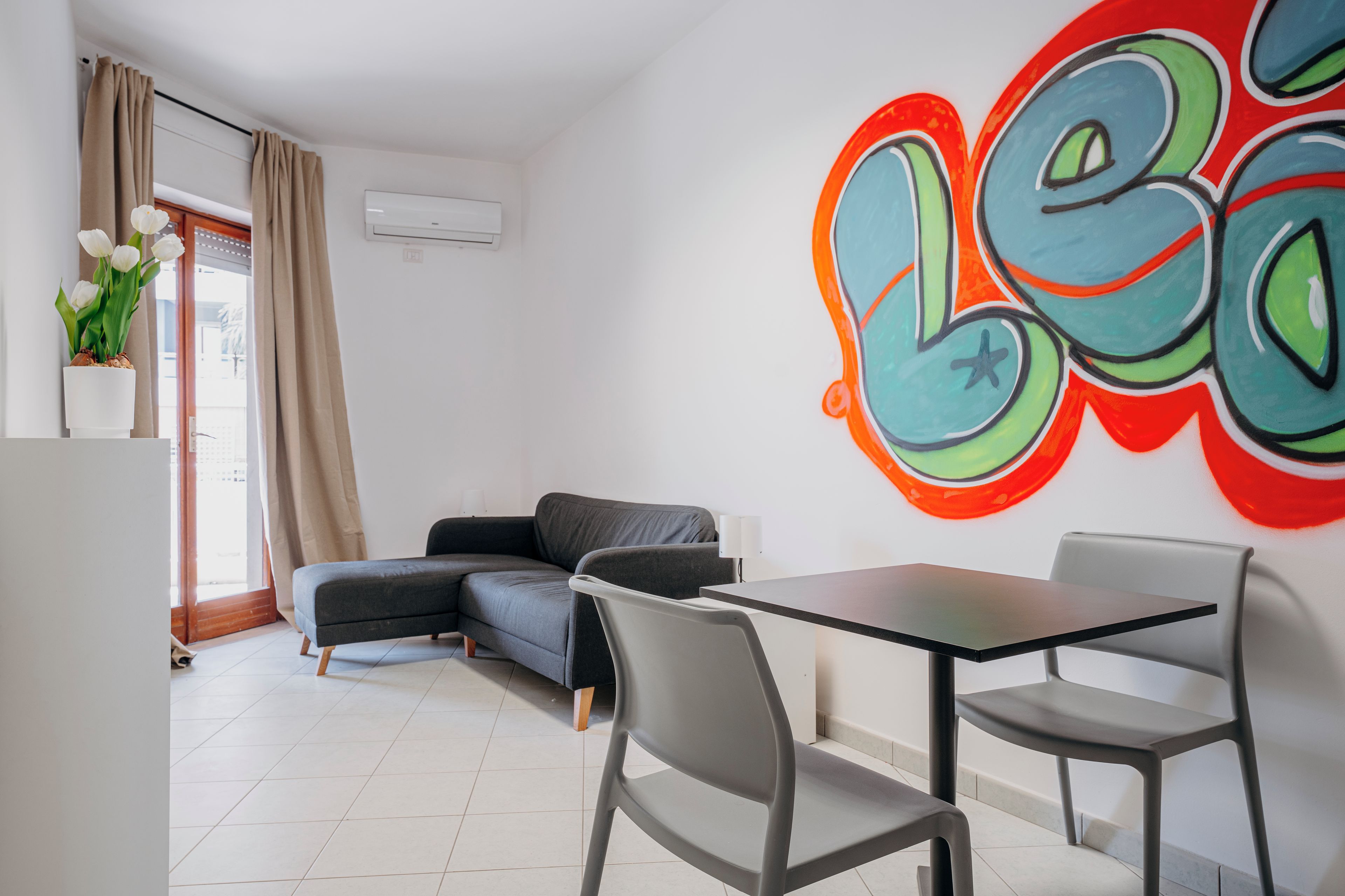 Una sala de estar moderna con un sofá gris, una mesa de comedor con sillas y un mural de pared colorido.