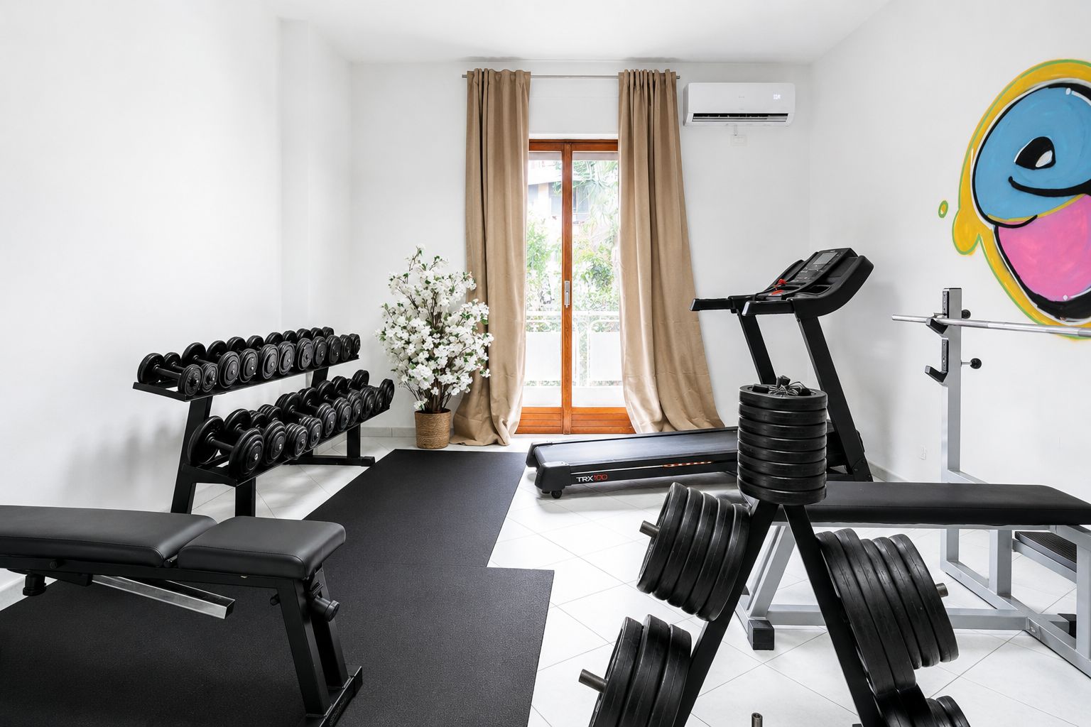 Ett gym med träningsutrustning, ett löpband och ett stort fönster med gardiner.