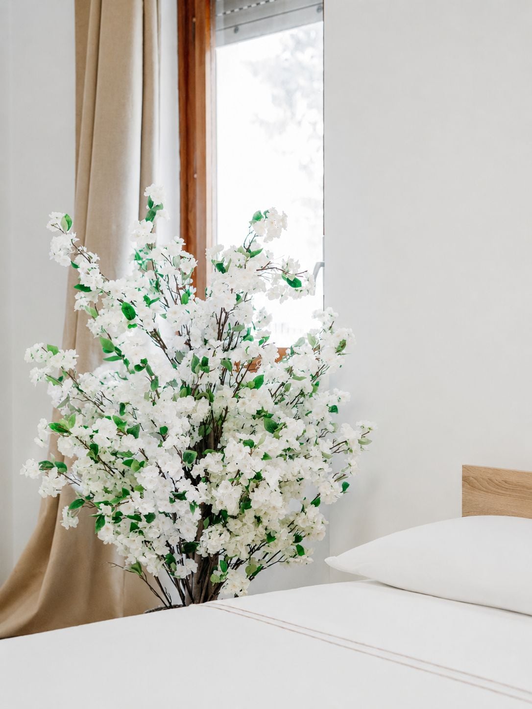 Ein Zimmer mit einem Bett, einem Vorhangfenster und einer Vase mit weißen Blumen.